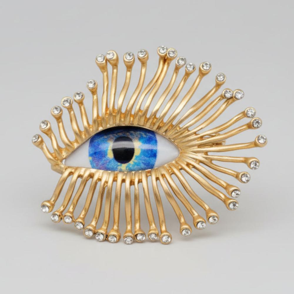 Decorative eye-shaped brooch with gold accents and gemstones on a light gray background